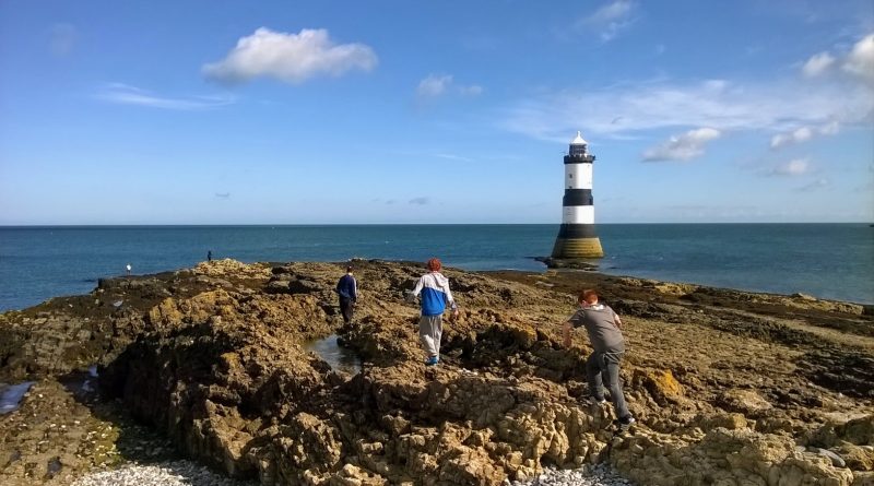 The image show children climbing over the rocks at Penmon Point heading towards the lighthouse